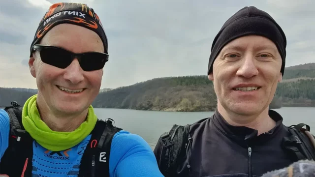Zwei Männer lächeln in die Kamera vor einem See